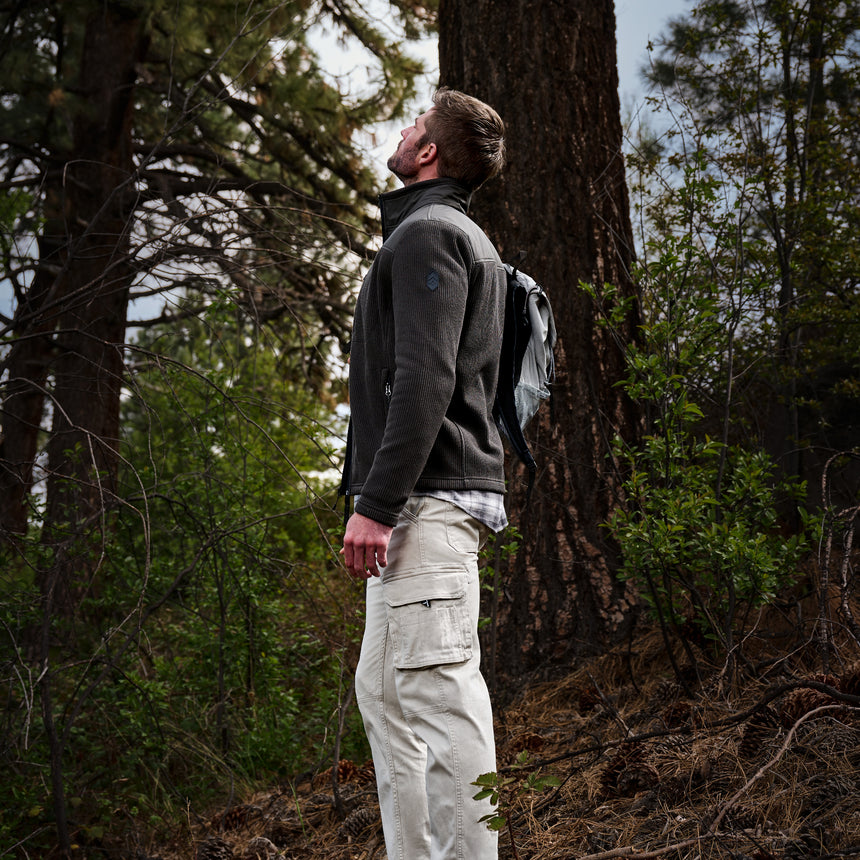 A man standing in a forest next to a large tree trunk, wearing a dark gray fleece jacket, light beige cargo pants, and a dark backpack.
