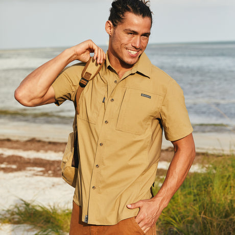 Man wearing the Free Country Men's Acadia Short Sleeve Shirt in Wheat, size M at beach, featuring a short sleeves, mesh underarm panels for breathability, and a zipper chest pocket made from lightweight polyester fabric, styled with orange shorts and backpack #color_ wheat
