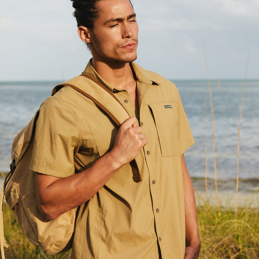 Man wearing the Free Country Men's Acadia Short Sleeve Shirt in Wheat, size M at beach, featuring a short sleeves, mesh underarm panels for breathability, and a zipper chest pocket made from lightweight polyester fabric, styled with orange shorts and backpack #color_ wheat