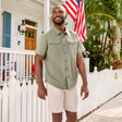 Man wearing Free Country Men's Explorer Short Sleeve Shirt in Sage, size M, made from lightweight quick drying polyester fabric featuring chest flap pockets, full button down front, and a stand collar, styled with white shorts in front of a beach house #color_sage