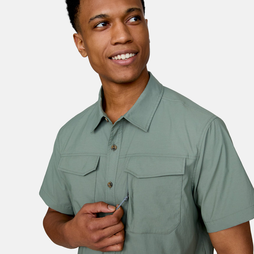 A person in the Clearance Men's Explorer Short Sleeve Shirt smiles as they unzip its small chest pocket. The shirt is lightweight with a button-down design, set against a plain light gray background.