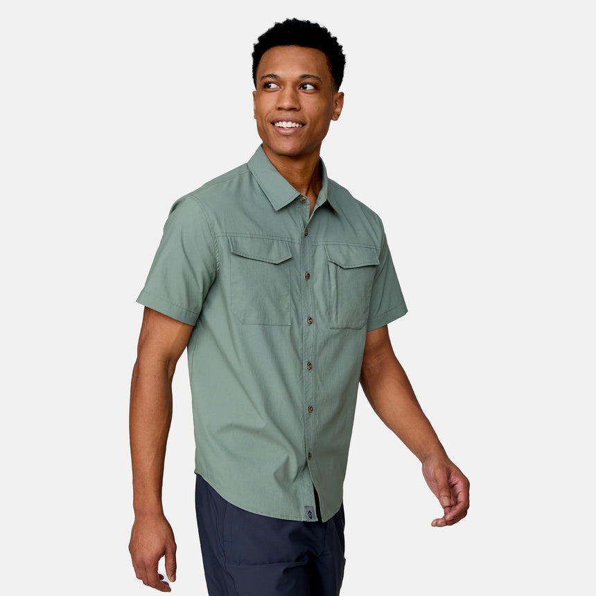 A man wearing the Clearance Men's Explorer Short Sleeve Shirt—a lightweight button-down with chest pockets—and dark pants stands against a plain light background, smiling and looking to the side.