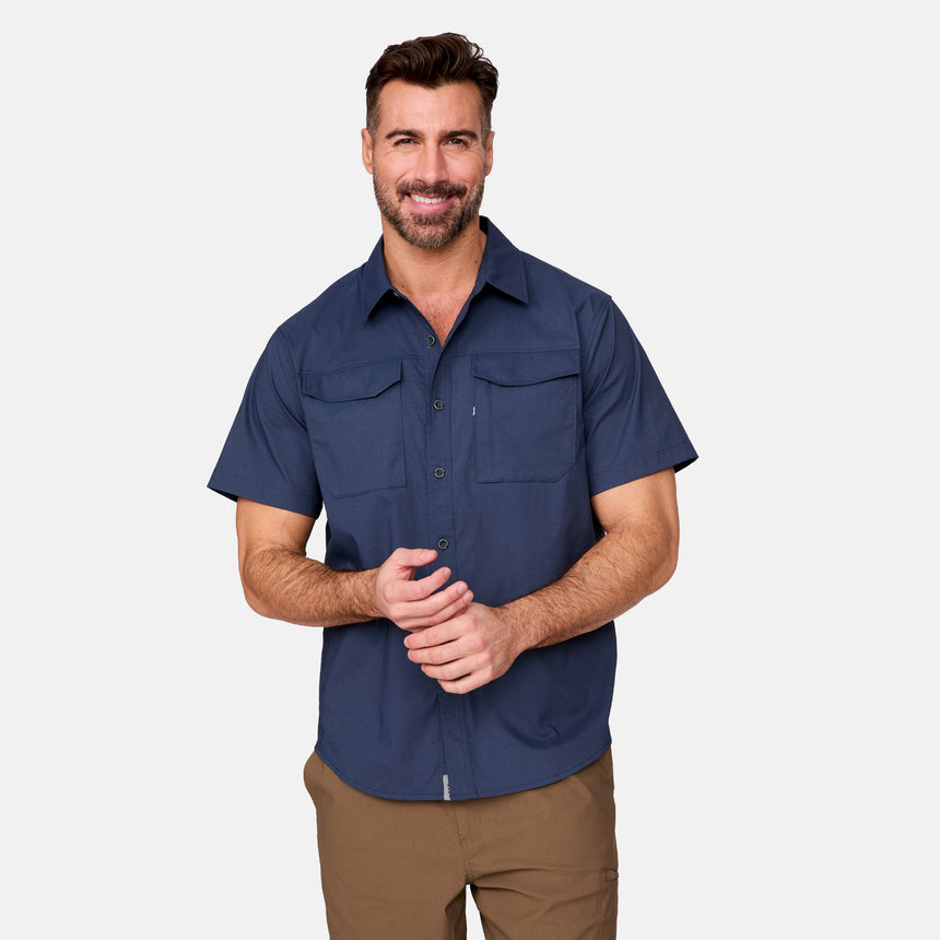 A man with short brown hair and a beard wears the Clearance Men's Explorer Short Sleeve Shirt, a lightweight button-down with two chest pockets, and brown pants, standing and smiling against a plain light grey background.