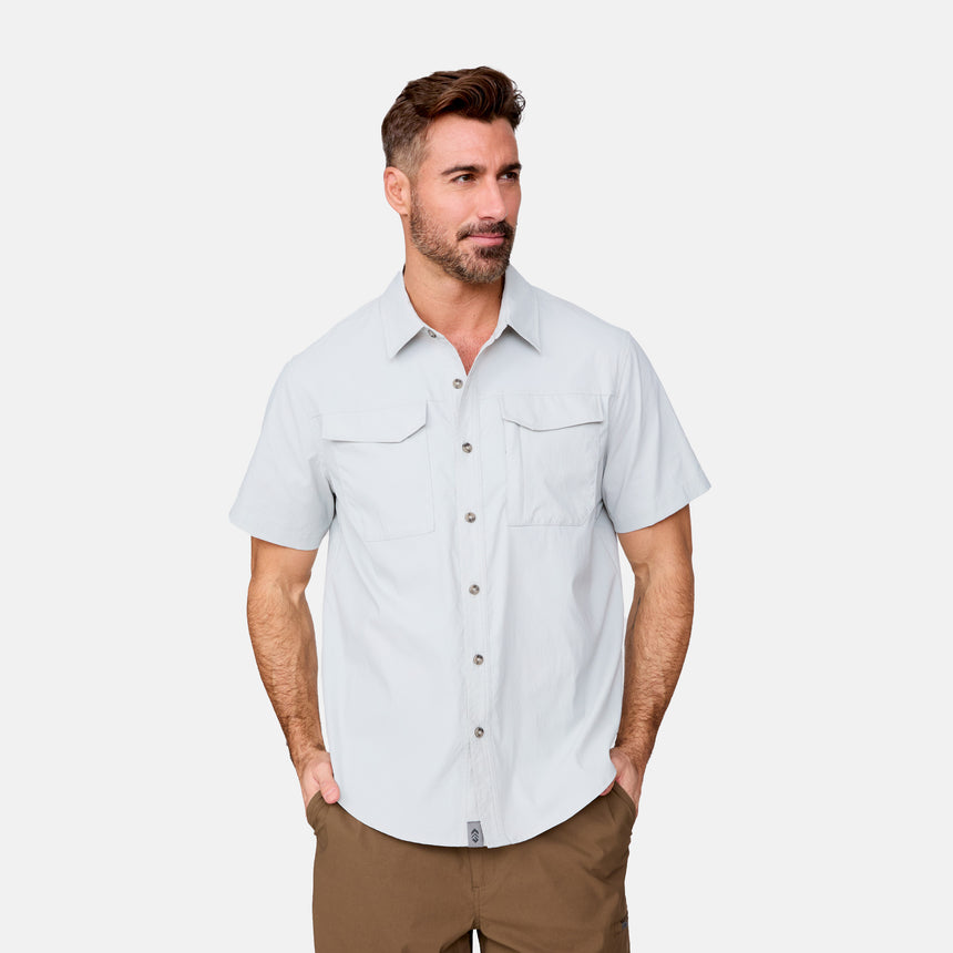 A man with short brown hair and a beard wears the Clearance Men's Explorer Short Sleeve Shirt, a lightweight button-down with two chest pockets, and brown pants, standing with his hands in his pockets against a plain white background.
