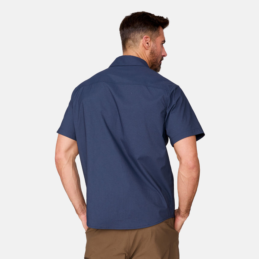 A man faces away with his hands in his pockets, wearing the Clearance Men's Explorer Short Sleeve Shirt in dark blue and brown pants. The lightweight button-down shirt has short sleeves and is set against a plain light background.