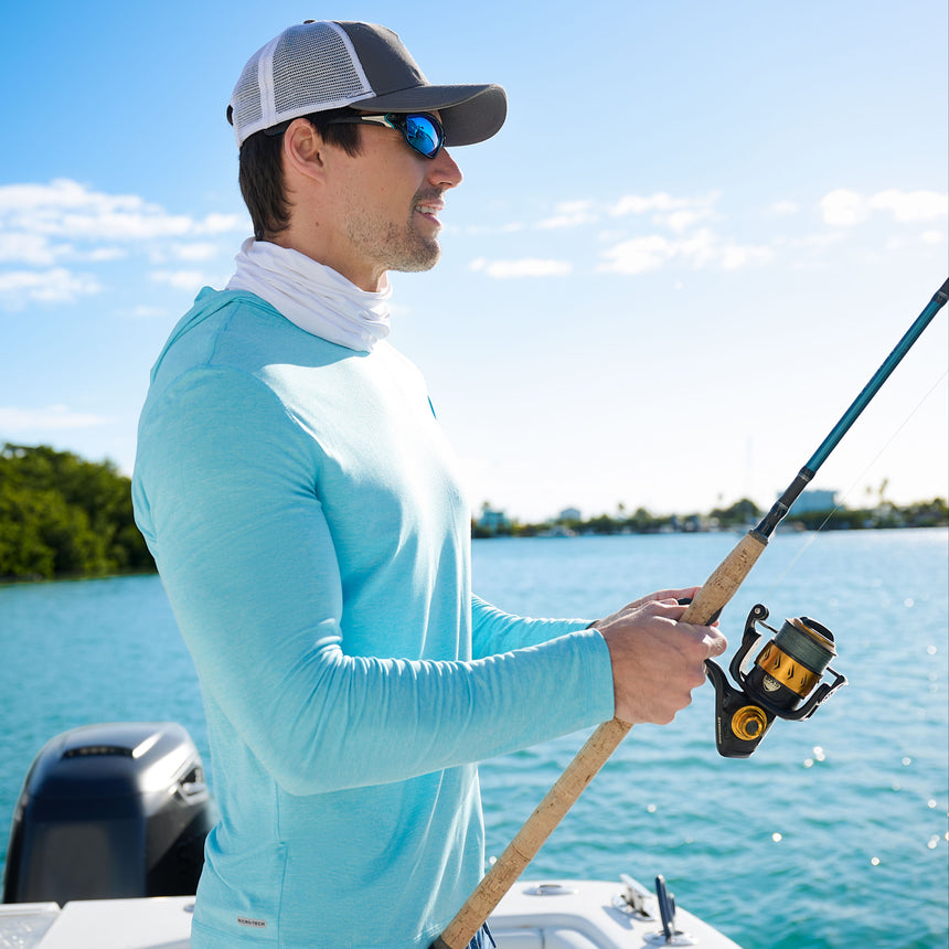A man wearing the Free Country Men's Microtech® Hooded Graphic Sunshirt, along with a cap, sunglasses, and neck gaiter, is fishing on a boat in sunny weather with blue water and trees in the background.