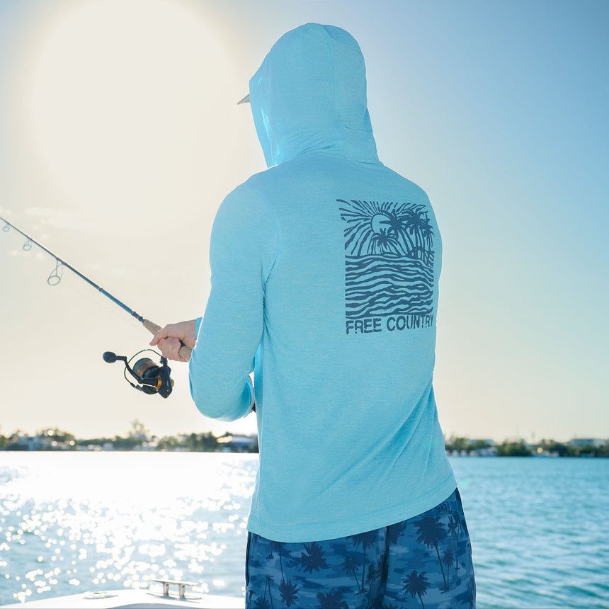 A man in a light blue Free Country Men's Microtech® Hooded Graphic Sunshirt with UPF 50+ and palm tree shorts fishes by calm water on a sunny day, "FREE COUNTRY" text and graphics displayed on the back.
