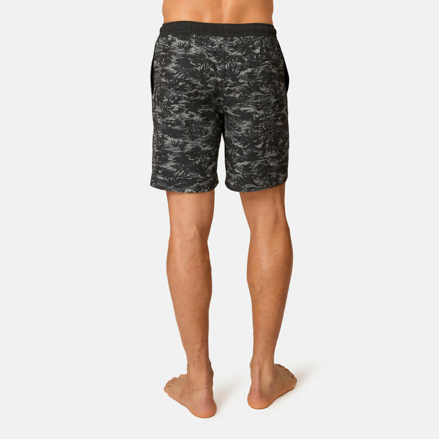 A person stands barefoot facing away, wearing Free Country Men's Surf Camo Swim Short with an elastic waistband. The black and gray camo swim shorts are shown from the waist down against a plain white background.