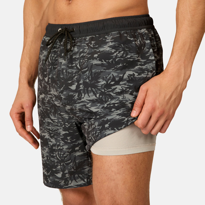 A man wears Free Country Men's Surf Camo Swim Shorts, featuring built-in light grey compression lining, lifting the outer camo layer to reveal the inner lining.