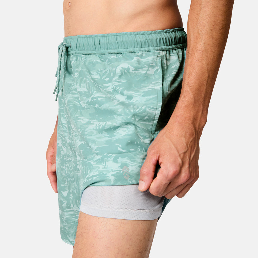 A man models Free Country's Men's Surf Camo Swim Short in green camo with a mesh lining, shown waist to mid-thigh against a plain background.