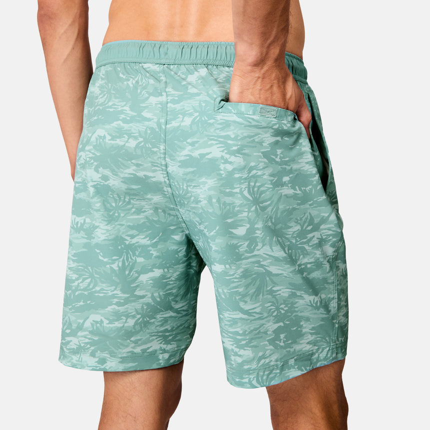 A person wears Free Country Men's Surf Camo Swim Short in light green with a leafy pattern, shown from the back with one hand in a pocket. These camo swim shorts offer an elastic waistband, relaxed fit, and UPF 50+ protection.