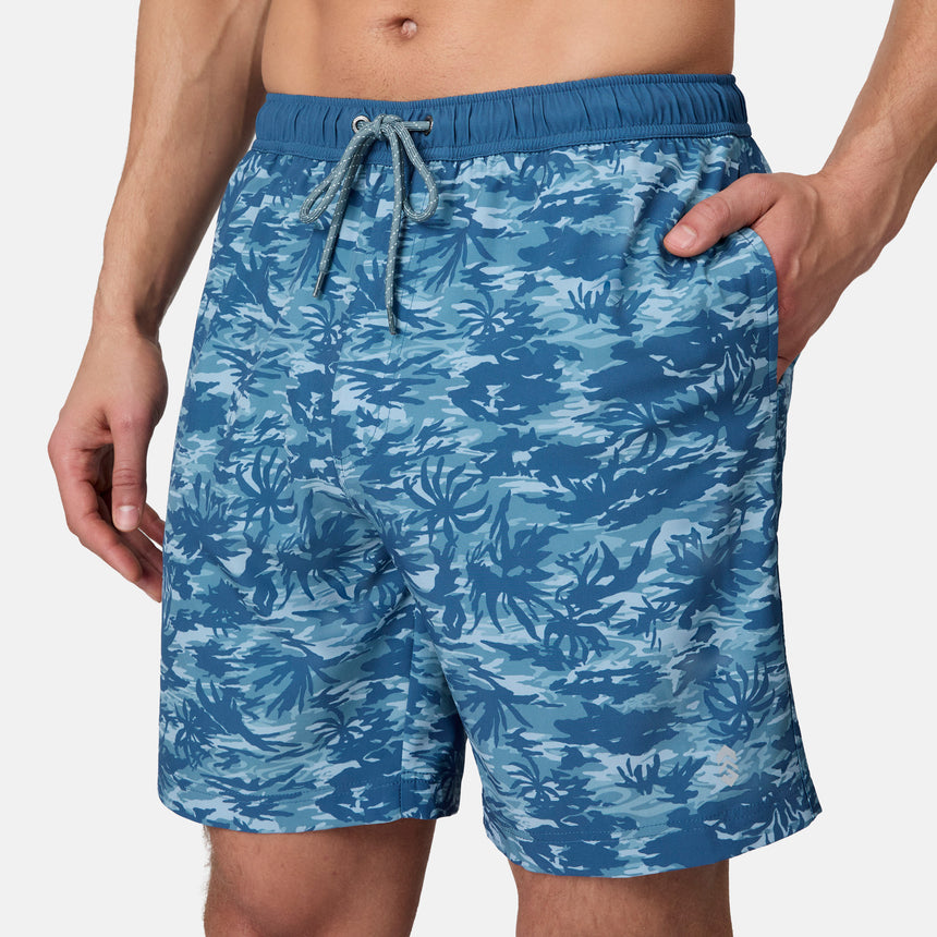 A man models the Free Country Men's Surf Camo Swim Short, featuring a blue camo pattern and drawstring waist, with one hand in his pocket. The swim shorts are shown cropped from mid-chest to mid-thigh.