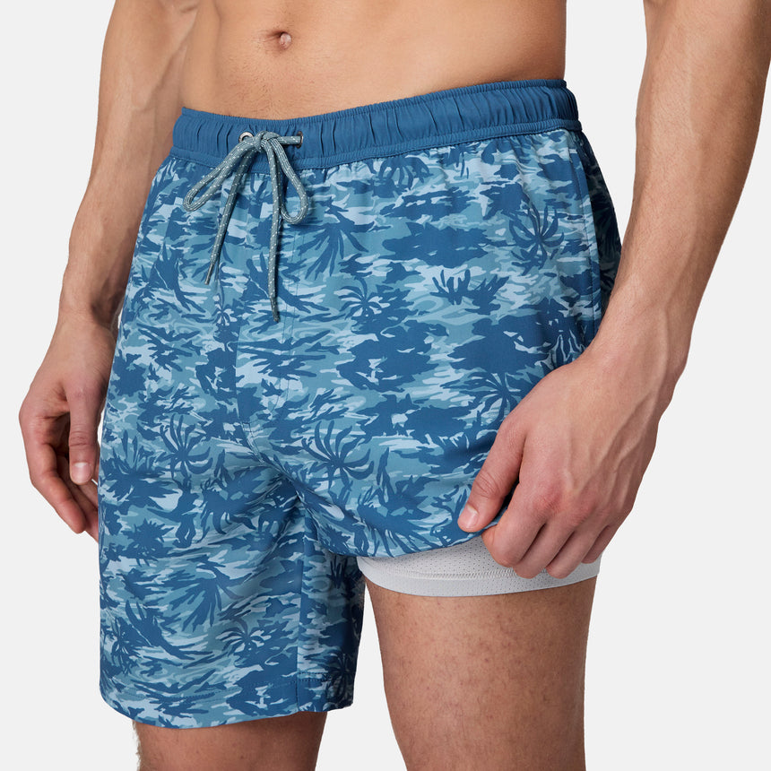 A person wears Free Country Men's Surf Camo Swim Shorts with a drawstring waistband and fitted white undershorts visible beneath, shown from waist to mid-thigh against a plain background.