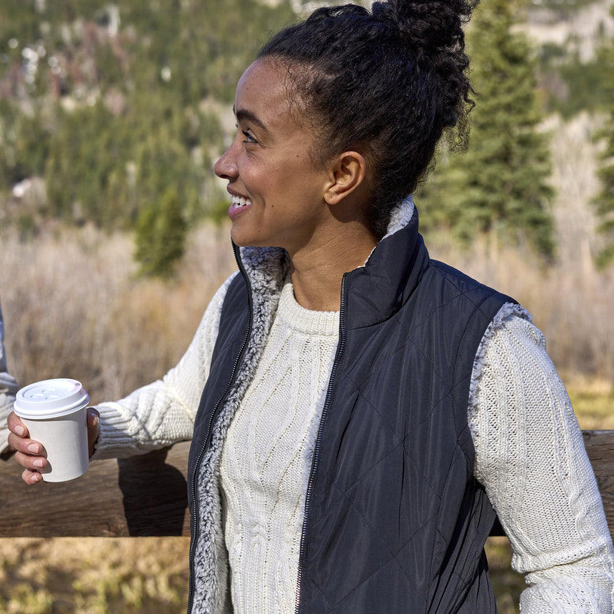 Woman wearing Free Country Women's Expedition Stratus Lite Reversible Vest in black with a full front zipper opening, soft lining, recycled polyester filling, and water resistant and wind resistant performance fabric. Best women’s vest for chilly weather.