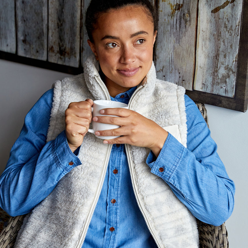 Woman wearing the Free Country Women's Venture Stratus Lite Reversible Vest in light gray, featuring a geometric-print Butter Pile® lining and horizontal quilted overlay, layered over a denim button-up shirt, holding a white mug indoors.