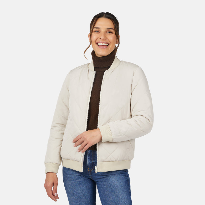 A smiling woman models the Clearance Women's Stratus Lite Reversible Quilted Bomber Jacket in cream over a dark brown turtleneck and blue jeans, standing against a plain white background.