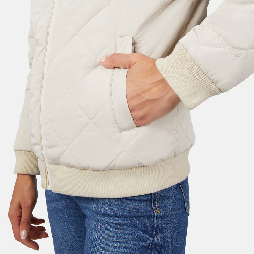 A person wears the Clearance Women's Stratus Lite Reversible Quilted Bomber Jacket in cream with ribbed cuffs and blue jeans, one hand in a pocket, standing against a plain white background.