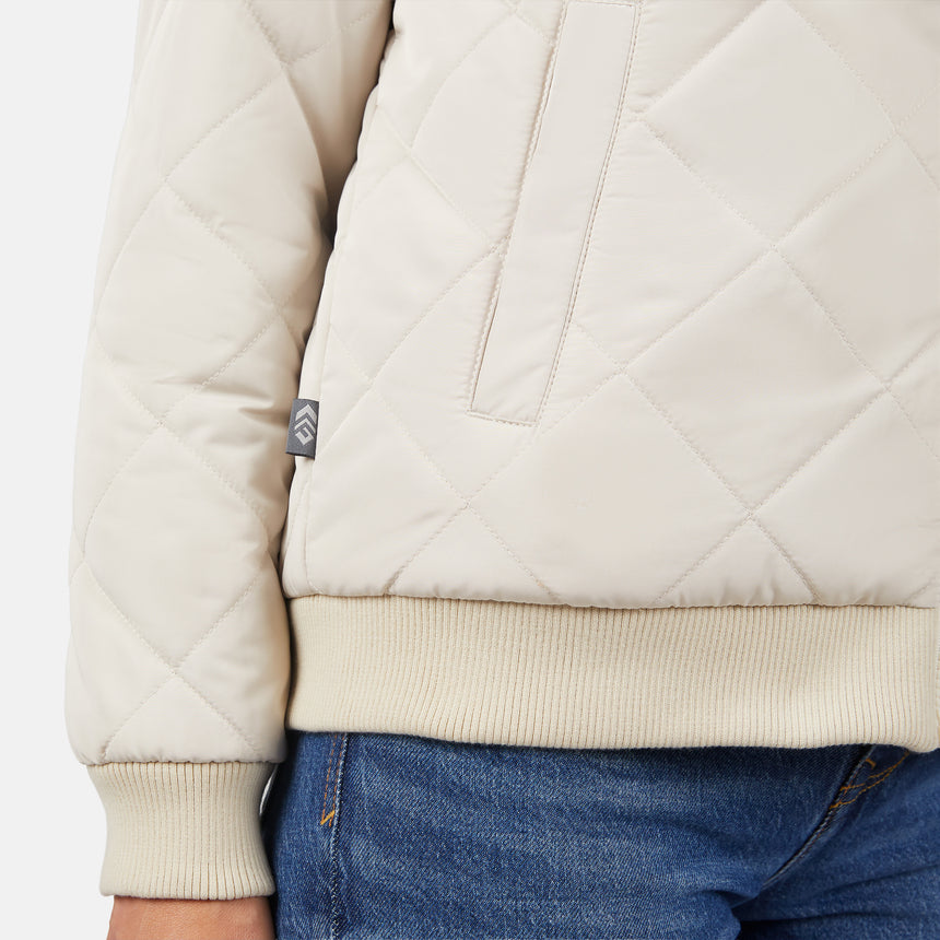 A close-up of a person wearing the Clearance Women's Stratus Lite Reversible Quilted Bomber Jacket in light beige with ribbed cuffs and hem, vertical pocket, small side tag, and blue denim jeans.