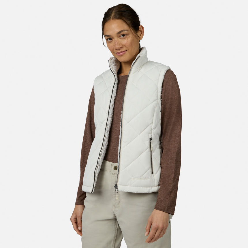 A woman stands against a plain white background, looking at the camera with a slight smile, wearing the Free Country Women’s Stratus Lite II Reversible Vest in white—crafted from recycled polyester—layered over a long-sleeve brown shirt and beige pants.