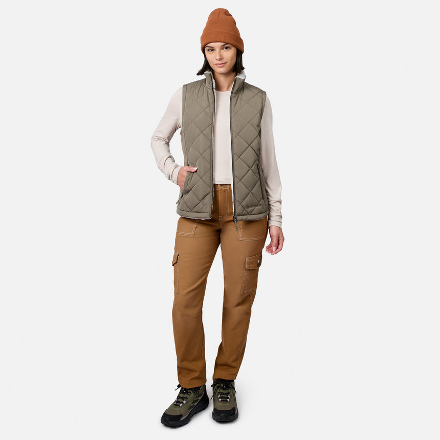 A person stands facing forward against a plain white background, wearing a brown beanie, the Free Country Women’s Stratus Lite II Reversible Vest—a quilted olive vest made from recycled polyester for lightweight warmth—a light long-sleeve shirt, brown cargo pants, and hiking boots.