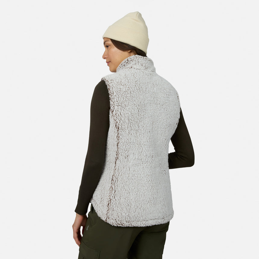 A woman stands facing away, looking over her shoulder against a plain white background while wearing a cream beanie, dark long-sleeve shirt, olive green pants, and the Free Country Women’s Stratus Lite II Reversible Vest in light gray sherpa fleece made from recycled polyester. This stylish vest delivers lightweight warmth with an eco-friendly touch.