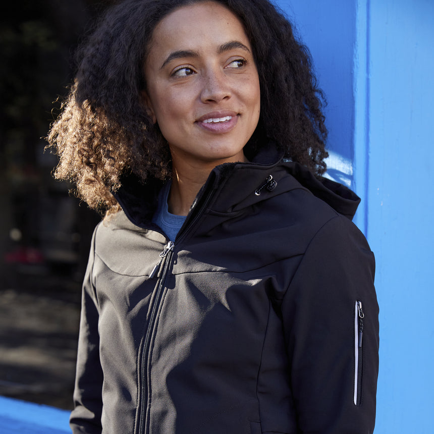 Woman wearing Free Country Women's StormTech Super Softshell® Jacket in black with a full front zipper opening, attached hood with adjustable cord, lower pockets, soft butter pile fabric lining, and water resistant and wind resistant stretch performance fabric. Perfect women’s jacket for fall weather.