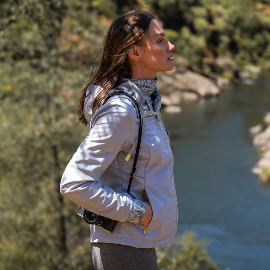 Woman wearing Free Country Women's Alps Super Softshell® Jacket in textured ligth grey with a full front zipper opening, soft lining, attached hood with adjustable drawcords, and water resistant and wind resistant stretch performance fabric, light grey background. Perfect women’s  jacket for fall weather.