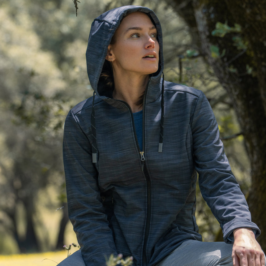 Woman wearing Free Country Women's Alps Super Softshell® Jacket in textured black with a full front zipper opening, soft lining, attached hood with adjustable drawcords, and water resistant and wind resistant stretch performance fabric, light grey background. Perfect women’s  jacket for fall weather.