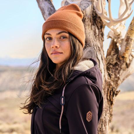 Woman wearing a brown beanie and purple jacket standing next to a tree in a desert-like setting