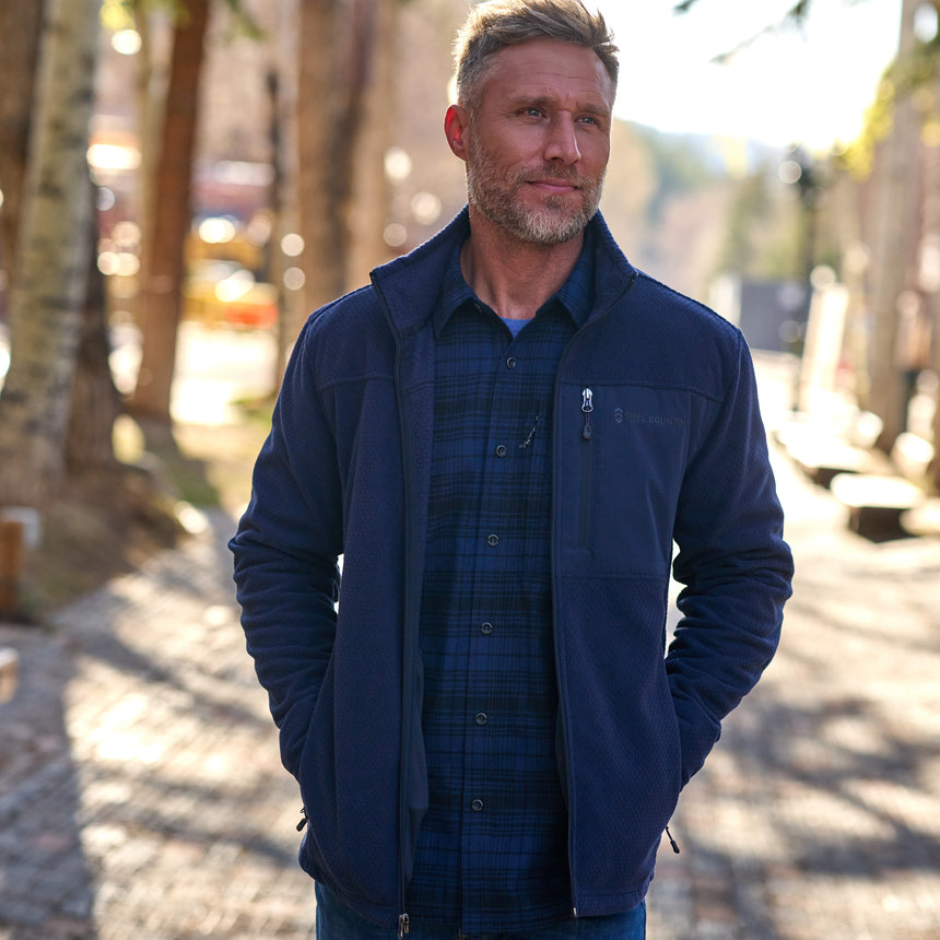 A man wearing a black fleece jacket with a full zip front closure, standing with hands in pockets.