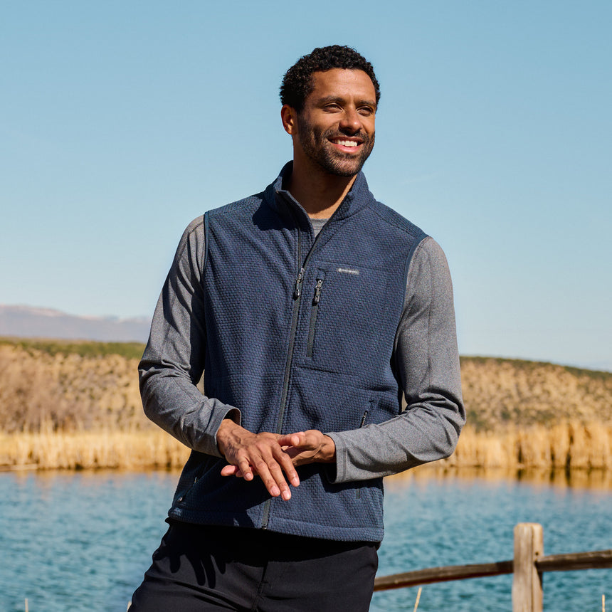 Man wearing a blue Free Country vest standing by a lake with mountains in the background