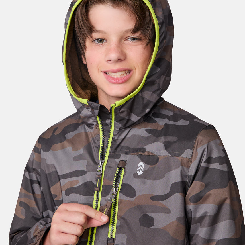 A young boy smiles wearing the Clearance Boys' Brisk Walk Windshear Jacket, featuring a camo print with a bright green zipper and hood lining.