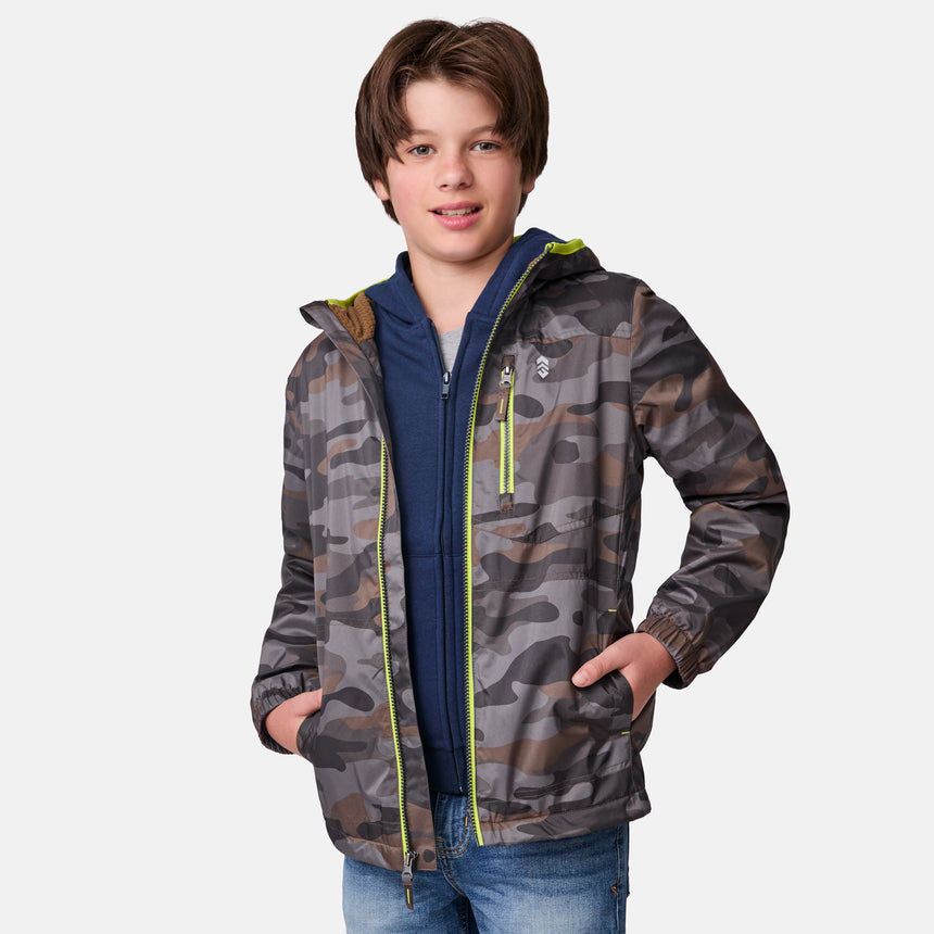 A boy with brown hair wears a Clearance Boys' Brisk Walk Windshear Jacket over a blue hoodie and jeans, standing with his hands in his jacket pockets and smiling at the camera against a plain white background.