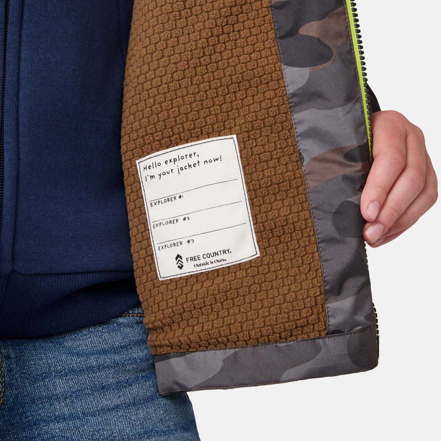A close-up of a person holding open the Boys' Brisk Walk Windshear Jacket by Clearance, featuring a camo print, brown textured lining, and a label saying, "Hello explorer, I'm your jacket now!" with spaces for explorer names.