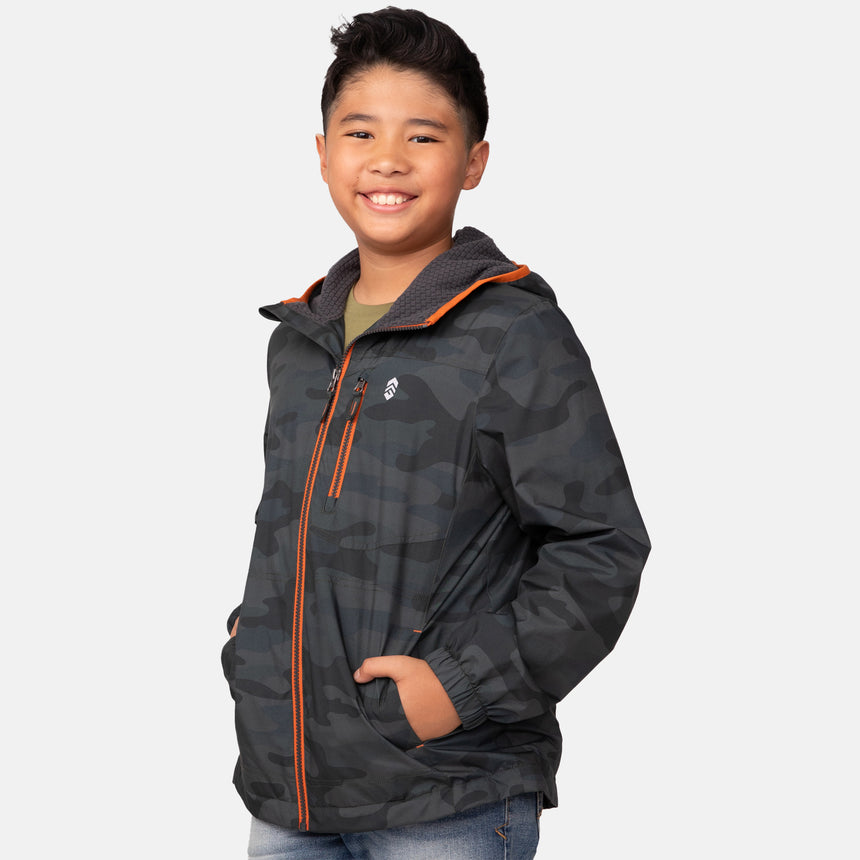 A smiling boy with short dark hair models the Clearance Boys' Brisk Walk Windshear Jacket in a dark camo print with orange zippers, hands in pockets, standing against a plain white background.
