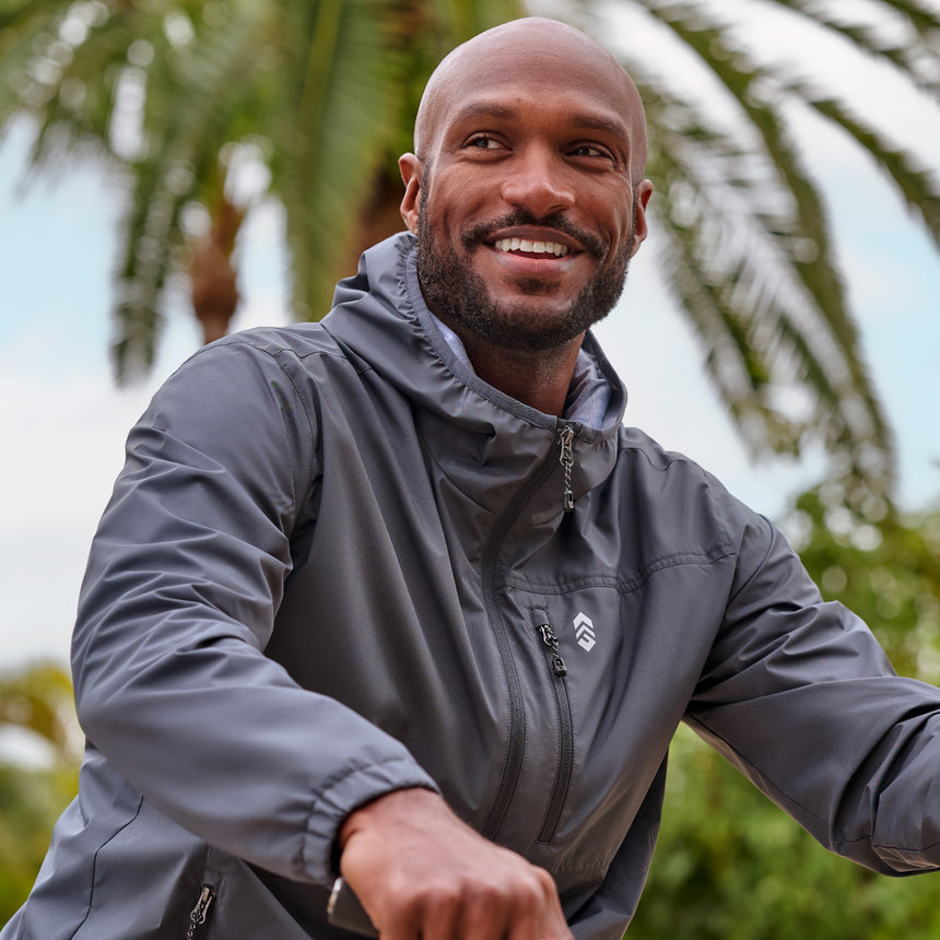 Man wearing the Free Country Men's Agile II Windshear Jacket in size M, made from lightweight wind and water resistant recycled polyester fabric and featuring an attached hood, inner phone pocket and elastic cuffs, styled with a black t shirt and black pants