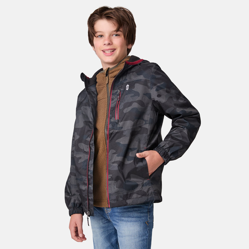 A smiling boy models the Clearance Boys' Brisk Walk Windshear Jacket in camo print over a brown zip-up sweater and blue jeans, standing against a plain white background.