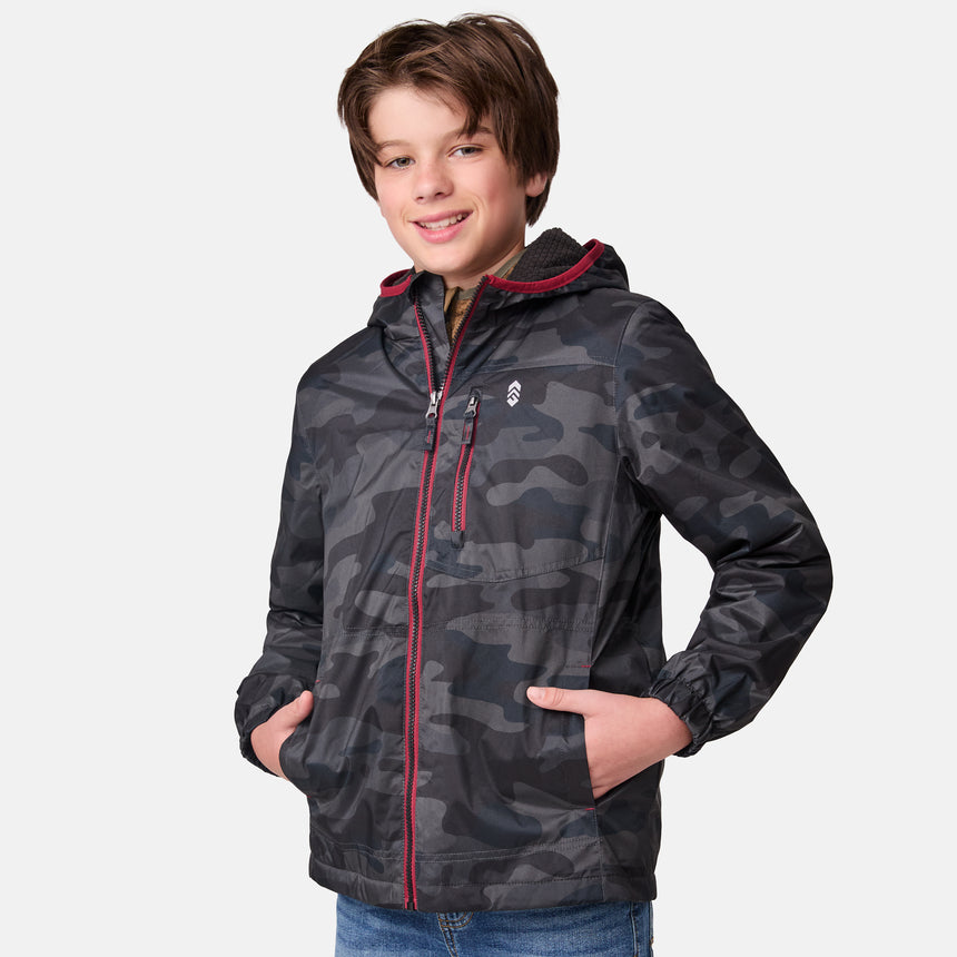 A young boy smiles at the camera in a Clearance Boys' Brisk Walk Windshear Jacket with a dark camo print and red zipper details, paired with blue jeans, standing against a plain white background.