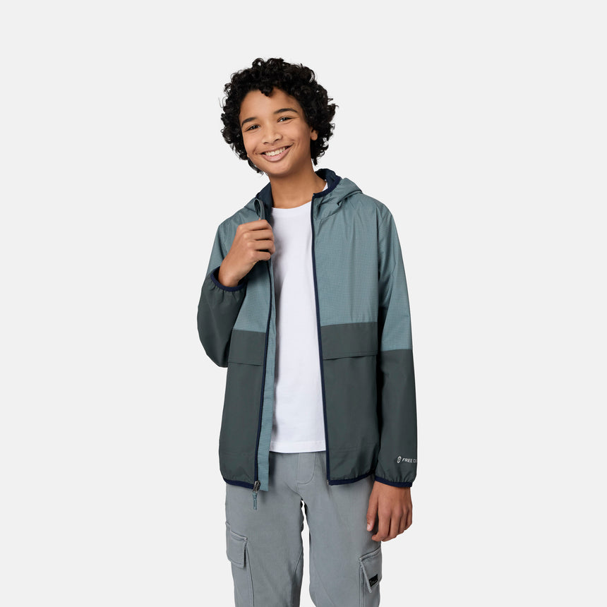A smiling teenager with curly hair wears the Free Country Boys' Easy Hiking Colorblock Windshear Jacket in teal and gray over a white t-shirt and gray pants, standing against a plain light gray background.