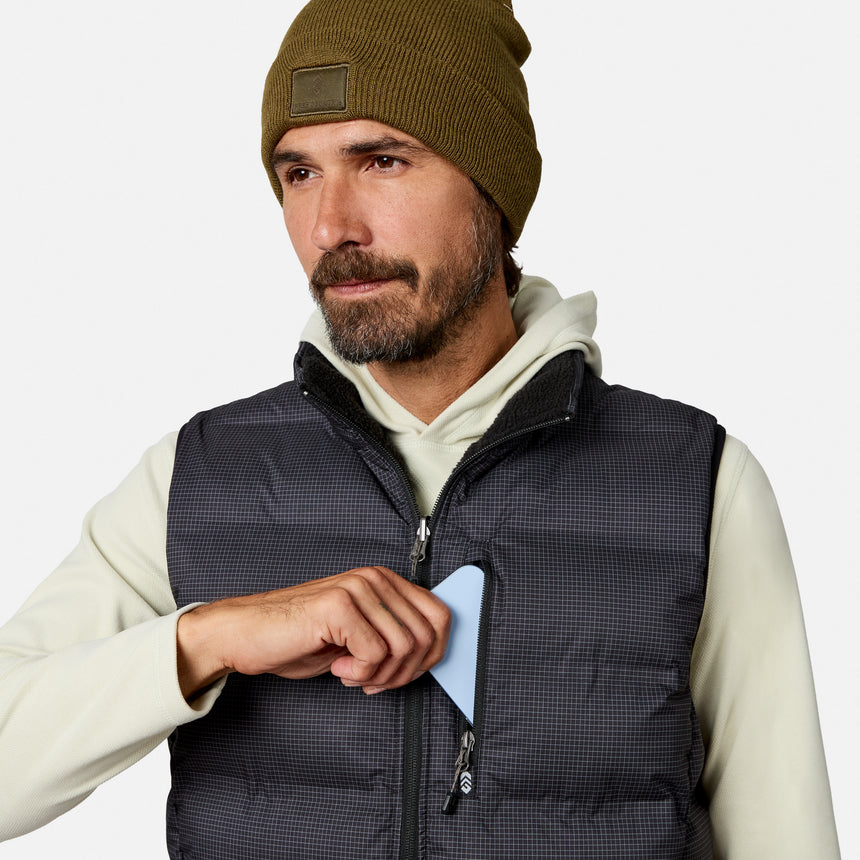 A man in a beanie and the Free Country Men’s Haven Stitchless Reversible Vest, renowned for its lightweight warmth, places a light blue object into the chest pocket of his vest. He looks to the side with a neutral expression against a plain background.