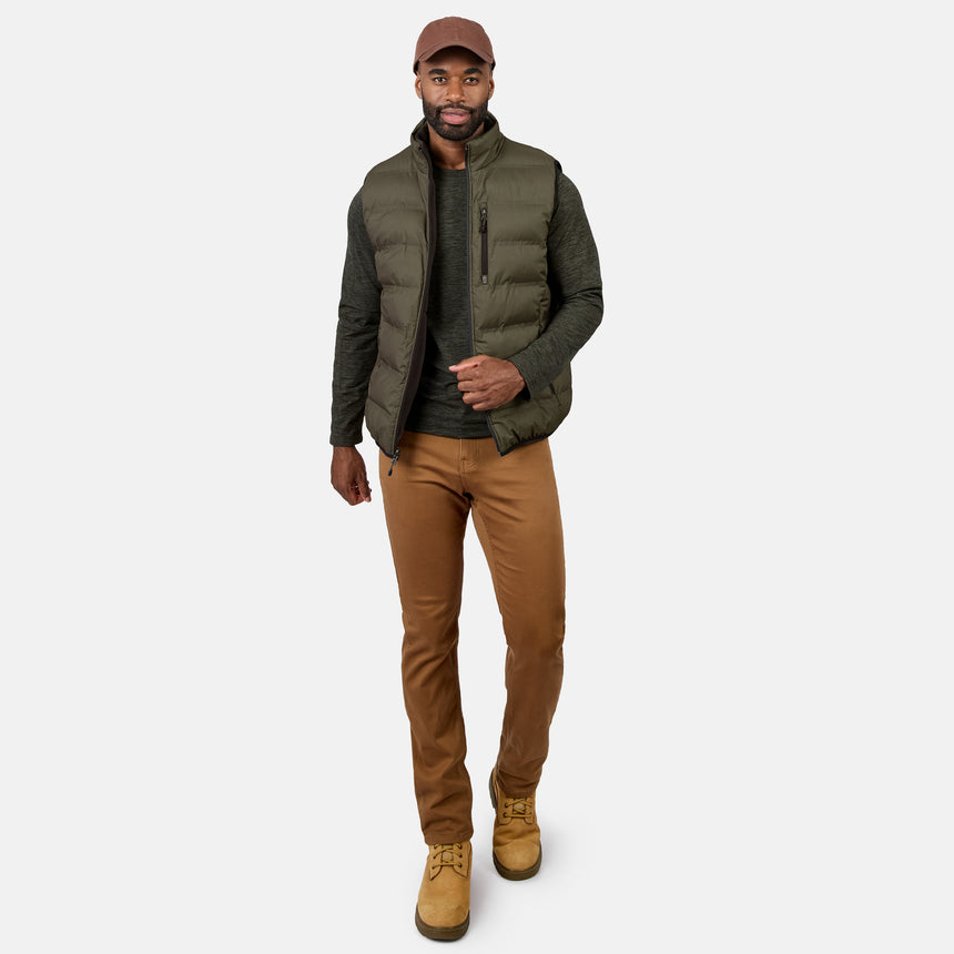 A man wearing a brown cap, the Free Country Men’s Haven Stitchless Reversible Vest for lightweight warmth in gray, a dark green long-sleeve shirt, tan pants, and tan boots stands against a plain white background, looking forward and walking confidently.