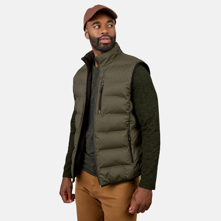 A man wearing a brown cap, an olive green long sleeve shirt, tan pants, and the Free Country Men’s Haven Stitchless Reversible Vest in Truffle stands against a light gray background, looking to the side.