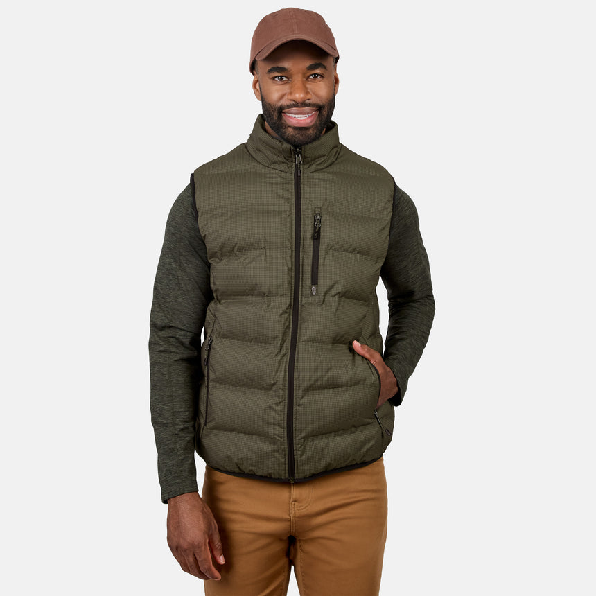 A man wearing the Free Country Men’s Haven Stitchless Reversible Vest in gray, along with a brown cap, green long-sleeve shirt, and tan pants, stands smiling with one hand in his pocket against a plain white background.