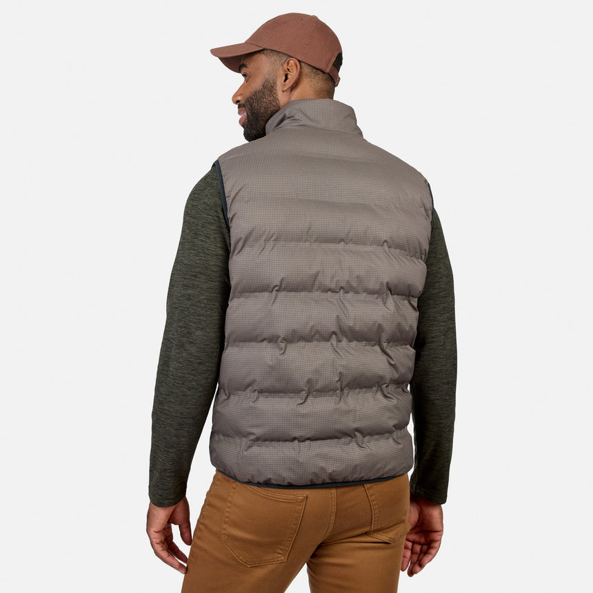 A man is standing and facing away from the camera against a plain white background, wearing a brown cap, green long-sleeve shirt, brown pants, and the Free Country Men’s Haven Stitchless Reversible Vest in gray, which provides lightweight warmth.