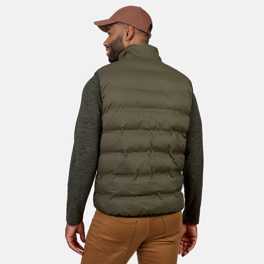 A man is standing and facing away from the camera against a plain white background, wearing a brown cap, green long-sleeve shirt, brown pants, and the Free Country Men’s Haven Stitchless Reversible Vest in gray, which provides lightweight warmth.