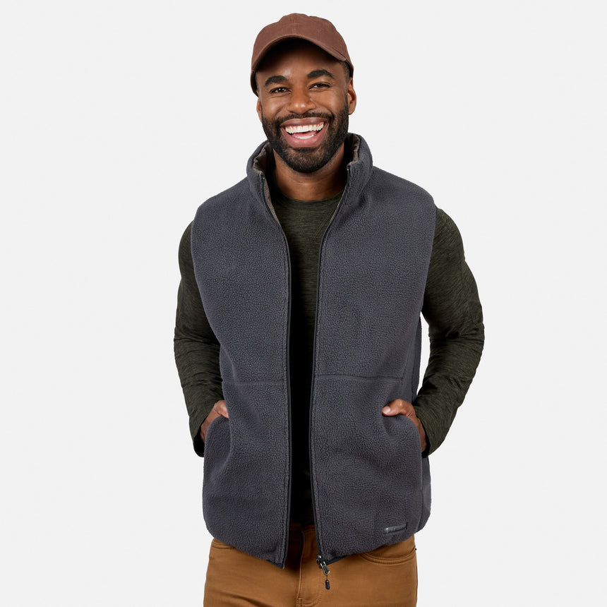 Smiling man wearing a brown cap, dark green long-sleeve shirt, brown pants, and the Free Country Men’s Haven Stitchless Reversible Vest in gray for lightweight warmth, standing with hands in vest pockets against a plain white background.