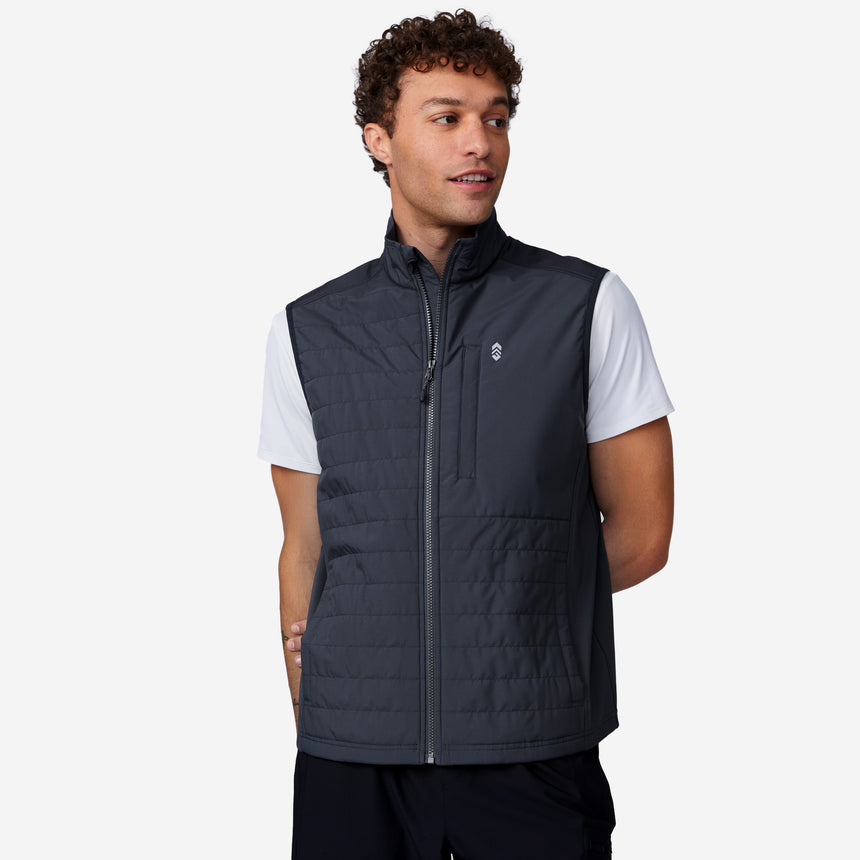 A young man models the Clearance Men's High Stretch Full Zip Vest in dark gray over a white short-sleeve shirt, standing against a light background and looking to his left with his left hand behind his back.