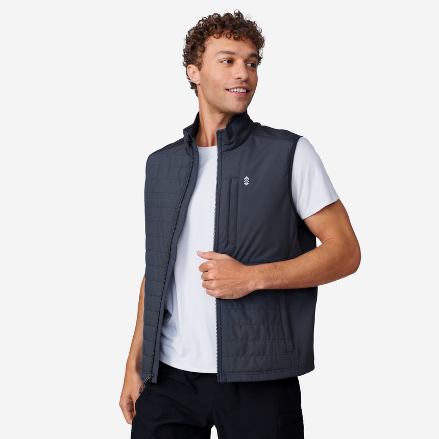 A man with curly hair smiles and looks left, wearing the Clearance Men's High Stretch Full Zip Vest in dark gray over a white t-shirt and black pants, standing against a plain light background.