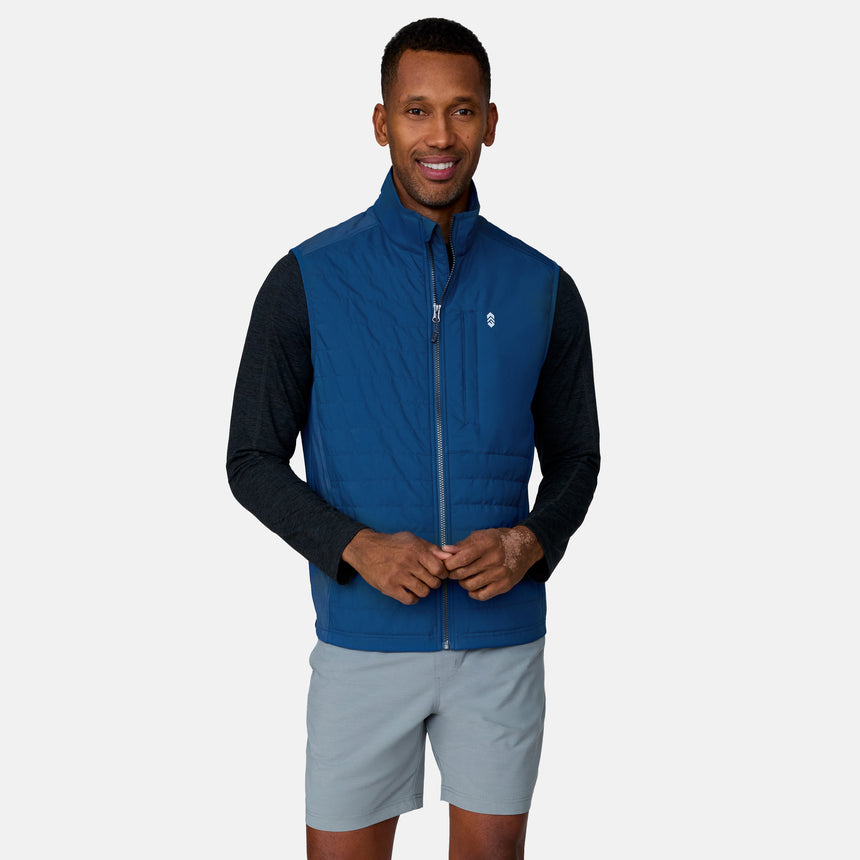 A man smiles against a plain light background, wearing the Clearance Men's High Stretch Full Zip Vest in blue over a dark long-sleeve shirt and light gray shorts.