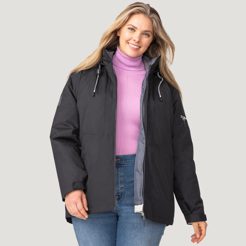 A woman wearing the Clearance Women's Plus Size Back of Bell 3-in-1 Systems Jacket in black over a lavender turtleneck sweater and blue jeans stands smiling against a plain light gray background.